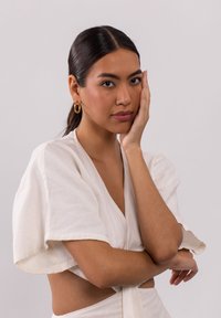 Model trägt ein Leinen-Oberteil in Cremefarbe mit weiten Ärmeln und einem geknoteten Akzent. Goldene Creolen sind sichtbar.