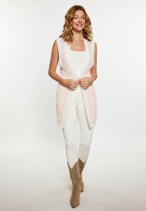 Langer, ärmelloser Cardigan mit einem hellrosa, strukturierten Muster und weißen Besätzen, kombiniert mit einem weißen Oberteil und cremefarbenen Hosen. Braune Stiefel mit Verzierung.