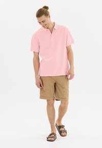 Ljusrosa polo skjorta med kort ärm, tillsammans med bruna shorts och bruna sandaler. Tyget verkar vara slätt med en klassisk kragsdesign.