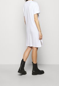 Biała bawełniana sukienka t-shirtowa z krótkimi rękawami i bocznymi rozcięciami, zestawiona z czarnymi, masywnymi botkami za kostkę. Prosty, casualowy design.