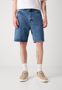 Denim shorts in medium blue met een relaxte pasvorm, een rafelzoom en een traditionele vijf-pocket ontwerp, gecombineerd met beige sneakers en witte sokken.