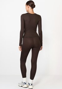 Barna, hosszúujjú felső és hozzáillő leggings, rugalmas anyagból készült. Feszes fazon jellemzi, finom textúrával. Fehér és kék sportcipővel viselve.