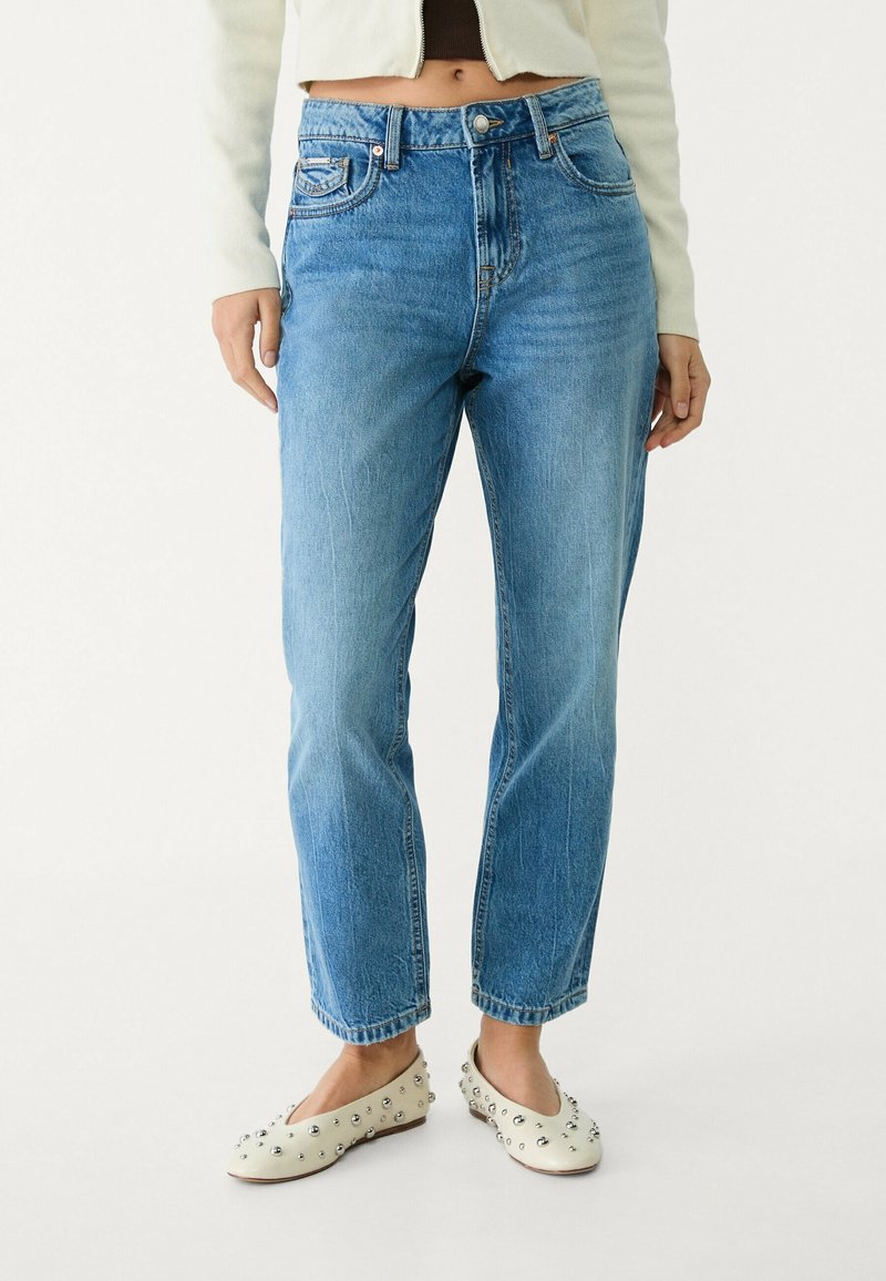 Femme portant un jean bleu clair taille haute, une veste blanche courte, un top noir et des ballerines blanches avec des clous argentés sur fond blanc.