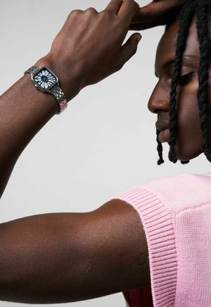 Montre bracelet carrée argentée avec un cadran floral bleu, attachée à un bracelet en métal texturé. Bras portant un haut en tricot rose dans une vue de profil rapprochée.