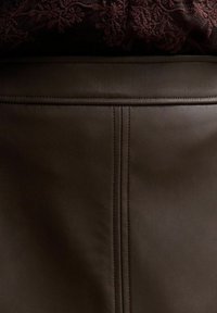 Abito in pelle marrone con cuciture visibili e una texture liscia, caratterizzato da un design a doppia cucitura in vita e dettagli floreali sopra.