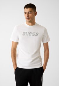 Weißes Baumwoll-T-Shirt mit einem strukturierten grauen "GUESS"-Logo. Kurzärmlig und mit Rundhalsausschnitt, kombiniert mit schwarzer Hose.