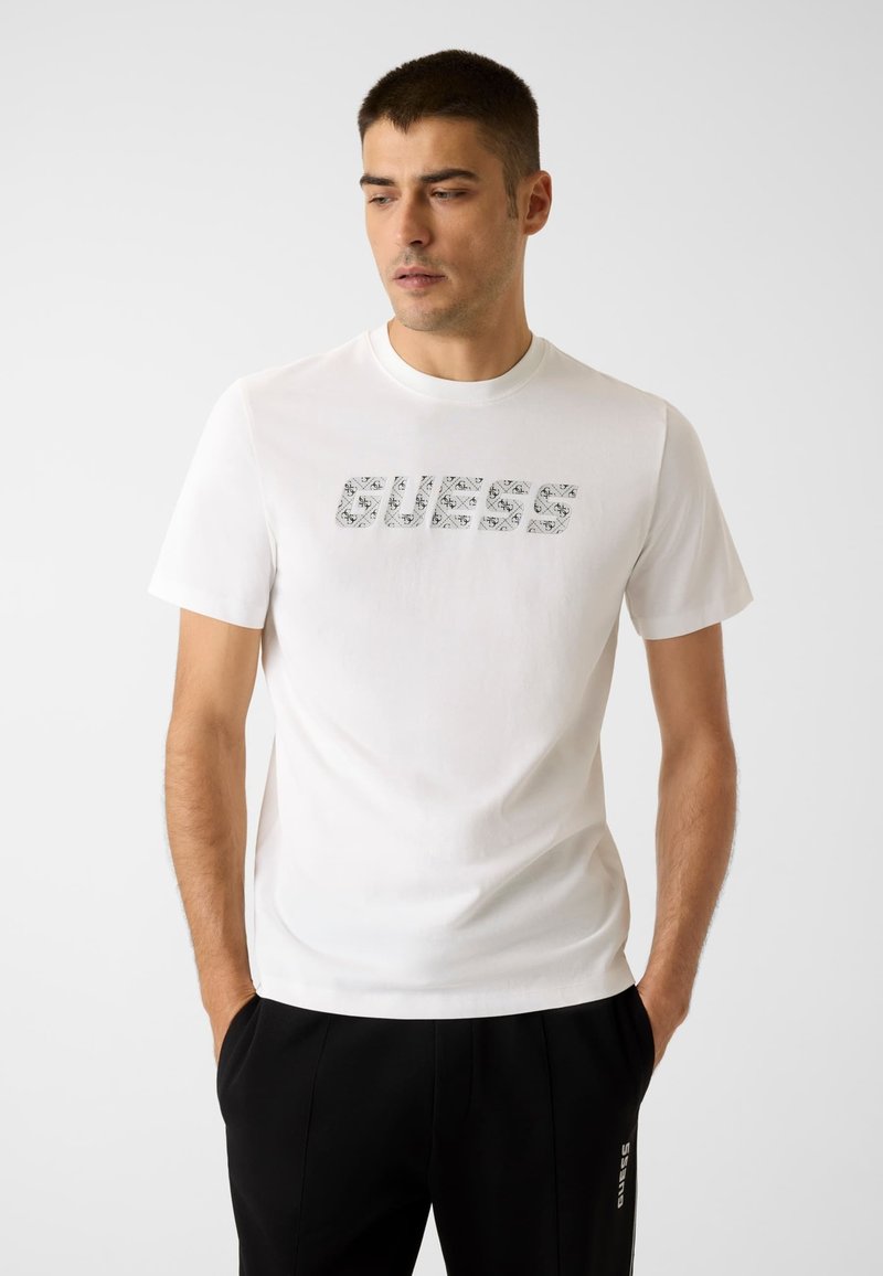 Weißes Baumwoll-T-Shirt mit einem strukturierten grauen "GUESS"-Logo. Kurzärmlig und mit Rundhalsausschnitt, kombiniert mit schwarzer Hose.