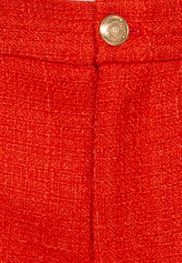 Červené tkané kalhoty s odpovídajícími pasem a výrazným zlatým knoflíkem na přední straně pro zapínání.