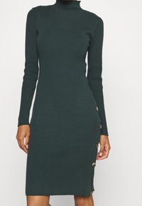 Ribbed turtleneck mekko tummassa vihreässä, jossa on pitkät hihat, istuva siluetti, sivupainikkeet ja polvipituinen leikkaus, jossa on sivu halkio.