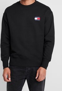 Felpa nera con collo a giro, polsini e orlo a coste. Presenta un piccolo logo rettangolare di Tommy Jeans in rosso, bianco e blu sul petto.