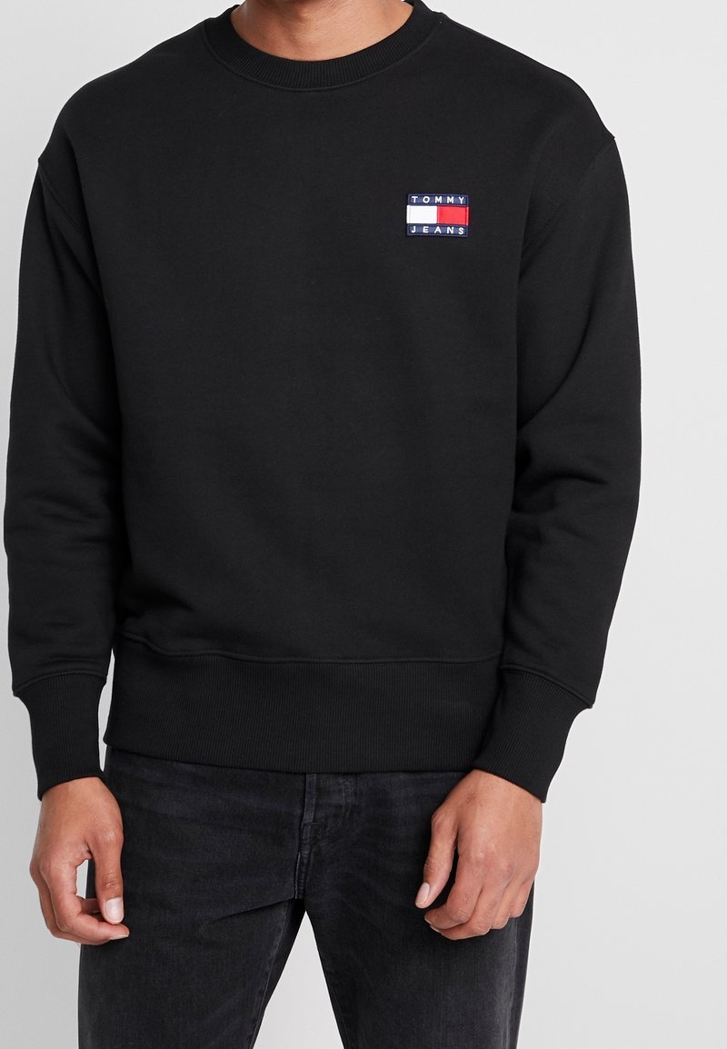 Felpa nera con collo a giro, polsini e orlo a coste. Presenta un piccolo logo rettangolare di Tommy Jeans in rosso, bianco e blu sul petto.