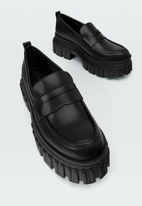 Mocassins en cuir noir avec une large sangle sur le dessus et des semelles en caoutchouc épaisses présentant des rainures et des crêtes.