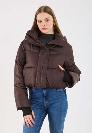 Bruine puffy gecropte jas met een hoge kraag, rits aan de voorkant en gestikte panelen. Gedragen over een zwart langemouwen top en blauwe jeans.