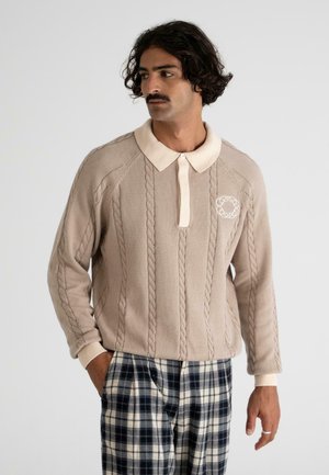 Beige knitted pullover with a textured cable pattern, featuring a cream-colored collar and a logo accent on the chest. Paired with plaid pants.