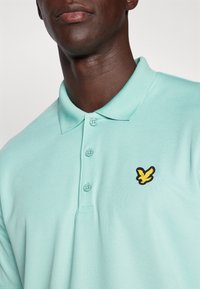 Lys blå polo skjorte laget av teksturert bomull med en tre-knaps placket og en svart og gul brodert logo på brystet.