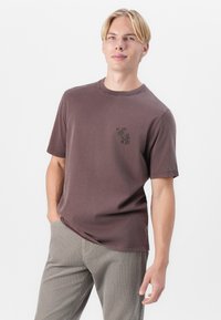 T-shirt en coton marron foncé avec un col rond et des manches courtes, ornée d'une petite impression graphique sur la poitrine.