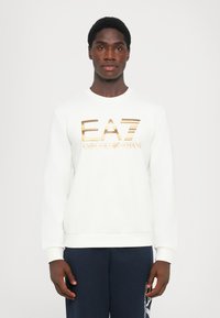 Sweatshirt branca feita de tecido macio com decote redondo. Apresenta um logótipo impresso em dourado brilhante, com punhos e barra canelados. Usada com calças azul-marinho.