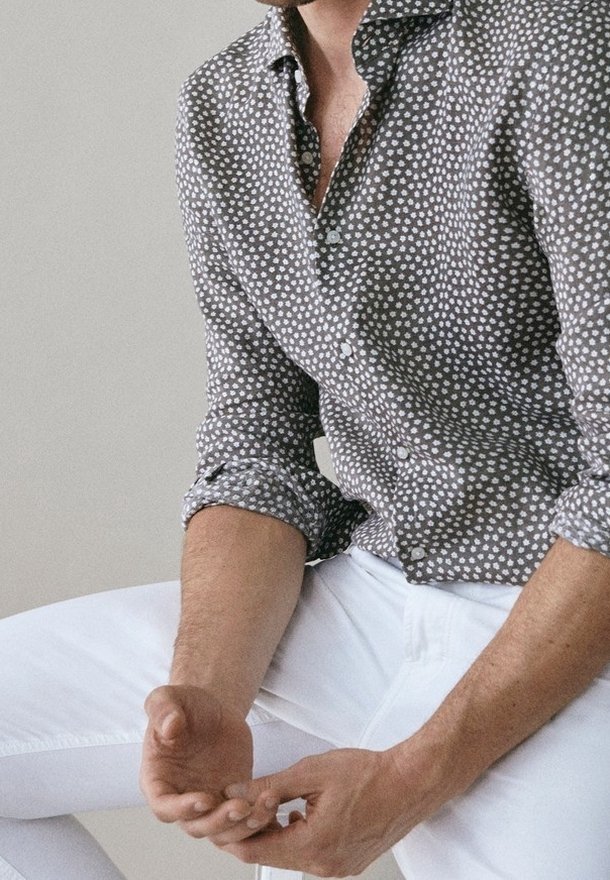 Homme assis portant une chemise grise à boutons avec un motif floral blanc et un pantalon blanc, mains jointes sur les genoux devant un fond uni.