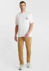 Wit katoenen T-shirt met grafische print; beige cargobroek met zijzakken; grijze sportschoenen; model staand tegen een effen achtergrond.