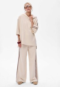 Vrouw in beige losse hoodie en brede broek met zijstrepen, met grote zonnebril, creolen, een armband en beige schoenen, staande.