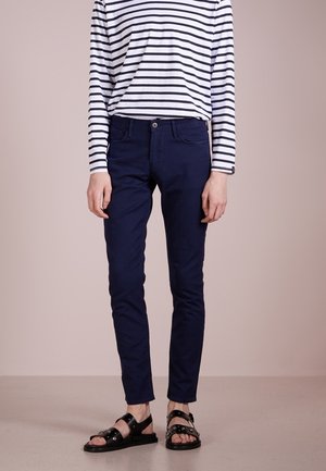 Person trägt marineblaue, schmal geschnittene Hosen, schwarze Sandalen mit Nieten und ein weißes Langarmshirt mit schwarzen horizontalen Streifen und steht auf einem neutralen Boden.