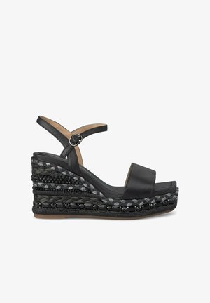 Sandale compensée noire avec une tige en cuir lisse, une sangle ajustable à la cheville et une plate-forme tressée texturée mettant en valeur des matériaux contrastants et des perles.