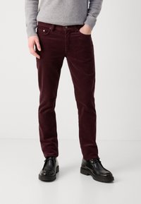 Pantalon en velours côtelé bordeaux à coupe slim, avec poches avant, fermeture à bouton, associé à des chaussures en cuir noires.