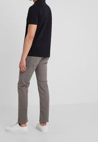 Hombre de pie contra un fondo liso, vistiendo una camiseta polo negra de manga corta, pantalones grises y zapatillas blancas, visto de lado.