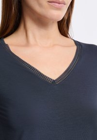 Mujer vistiendo una blusa azul marino con cuello en V y delicados bordados de encaje en el escote, mostrando parte del cuello y los hombros.