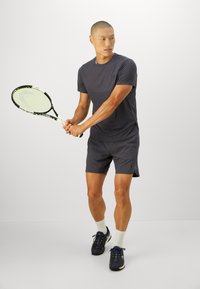 Mand i mørkt sportsbeklædning, der holder en tennisracket med begge hænder, forbereder sig på at slå til en bold, stående på en ensfarvet hvid baggrund.
