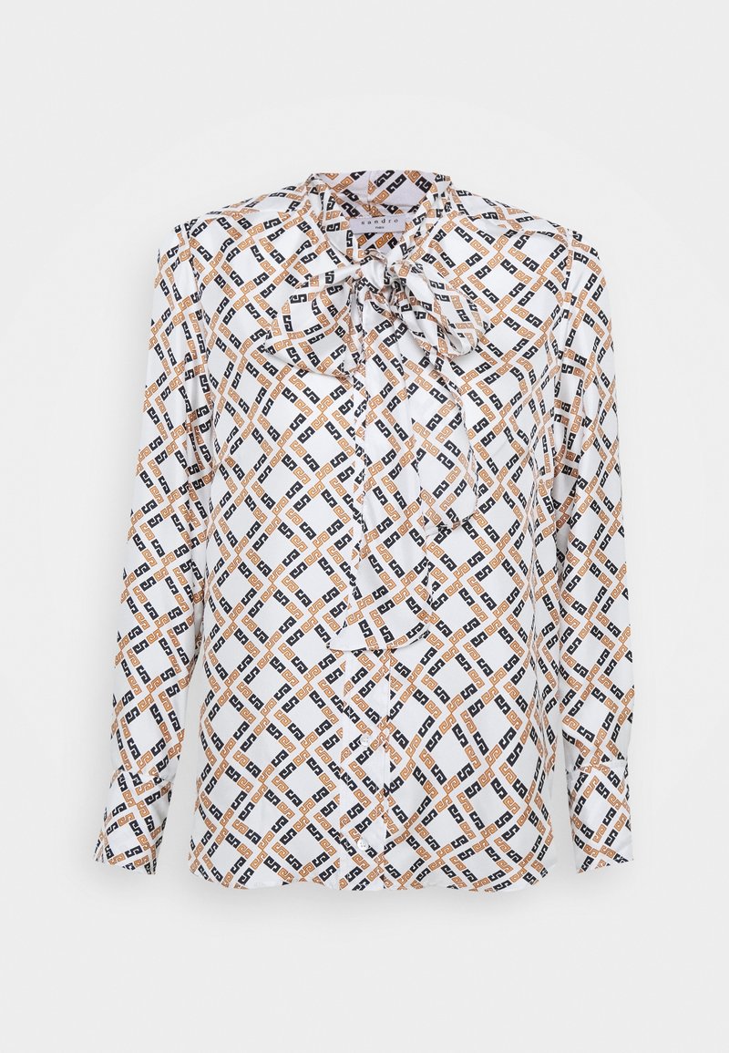 Blouse blanche à manches longues avec motif géométrique en chaînes noires et orange et un nœud noué au col, présentée sur un fond uni.