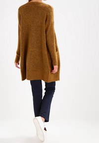 Suéter marrón de punto con mangas largas, corte holgado y escote redondo, combinado con pantalones oscuros y zapatillas blancas.