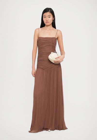 Brown, ruched maxi dress with spaghetti straps, flowing skirt, and a lightweight texture; paired with a small, cream clutch bag.