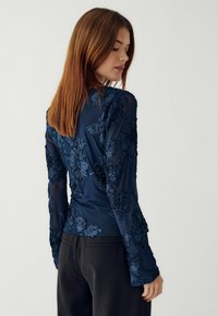 Jūras spēku ilgas piedurknes tops ar ziedu izšuvumiem. Izgatavots no viegla auduma, ar pieguļošu dizainu un teksturētām akcentiem uz piedurknēm.