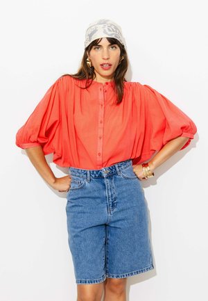 Femme portant un chemisier corail à manches bouffantes, un short en jean taille haute, un foulard à motifs et des bracelets en or, debout les mains sur les hanches.