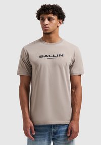 Beige T-shirt met korte mouwen van een zachte stof, met een ronde hals en een zwarte "BALLIN'"-logo op de borst. Draagt bij een spijkerbroek.
