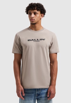 Beige T-shirt met korte mouwen van een zachte stof, met een ronde hals en een zwarte "BALLIN'"-logo op de borst. Draagt bij een spijkerbroek.