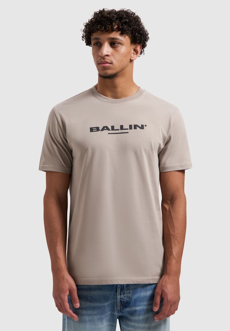 Beige T-shirt met korte mouwen van een zachte stof, met een ronde hals en een zwarte "BALLIN'"-logo op de borst. Draagt bij een spijkerbroek.