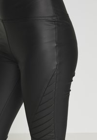 Svarta faux läderleggings med hög midja och diagonala sömnadsdetaljer på sidorna, som visar en slank och figurnära design.