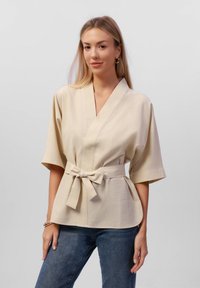 Top avvolgente beige con maniche corte, con cintura in vita, realizzato in tessuto testurizzato, caratterizzato da un scollo a V e un orlo curvo.