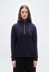 Νεαρή γυναίκα με μακριά καστανά μαλλιά φοράει ένα μπλε σκούρο φούτερ με φερμουάρ μέχρι τη μέση και ένα μικρό λογότυπο στο στήθος, στέκεται μπροστά σε ένα απλό λευκό φόντο.