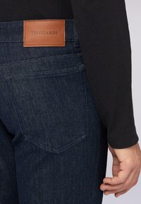 Pantalones vaqueros de denim azul oscuro con un parche de cuero etiquetado "TRUSSARDI" en la parte posterior. La tela tiene una textura suave con sutiles detalles de costura.