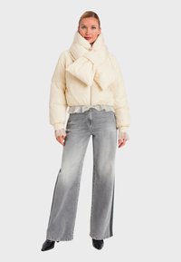 Giaccone imbottito color crema con collo a fiocco oversize, dotato di cerniera frontale. Abbinato a jeans grigi a gamba larga e stivaletti neri alla caviglia.