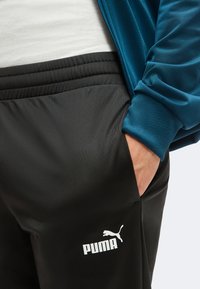 Άτομο με μαύρο παντελόνι φόρμας Puma και μπλε μπουφάν με το ένα χέρι στην τσέπη, δείχνοντας το λογότυπο Puma στο παντελόνι.