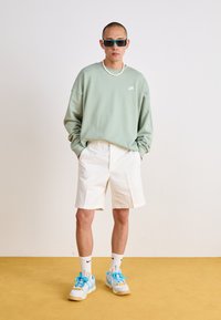 Felpa oversize verde menta, pantaloni corti bianchi e sneakers con dettagli blu. Gli accessori includono occhiali da sole e una collana di perline.
