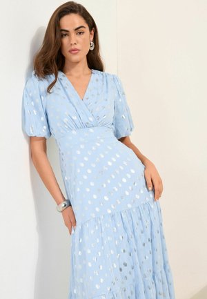 Nő hosszú barna hajjal, ezüst karika fülbevalóval és világoskék ruhával, ezüst pöttyökkel, fehér falnak támaszkodva.