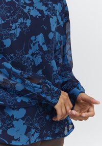 Blusa trasparente blu navy con un motivo floreale in blu chiaro. Presenta maniche lunghe con polsini bottonati e una vestibilità ampia e fluida.