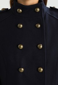 Abrigo de lana azul marino con un alto cuello, que presenta seis botones dorados decorativos y distintivos epaulets en los hombros.