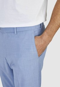 Lichtblauwe broek van gestructureerde stof met een vlakke voorkant, zijzakken en een slim fit. Een hand rust in de rechterzak.