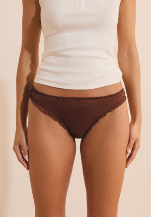 Mujer con top blanco sin mangas y ropa interior estilo bikini marrón, de pie con las manos relajadas a los lados.